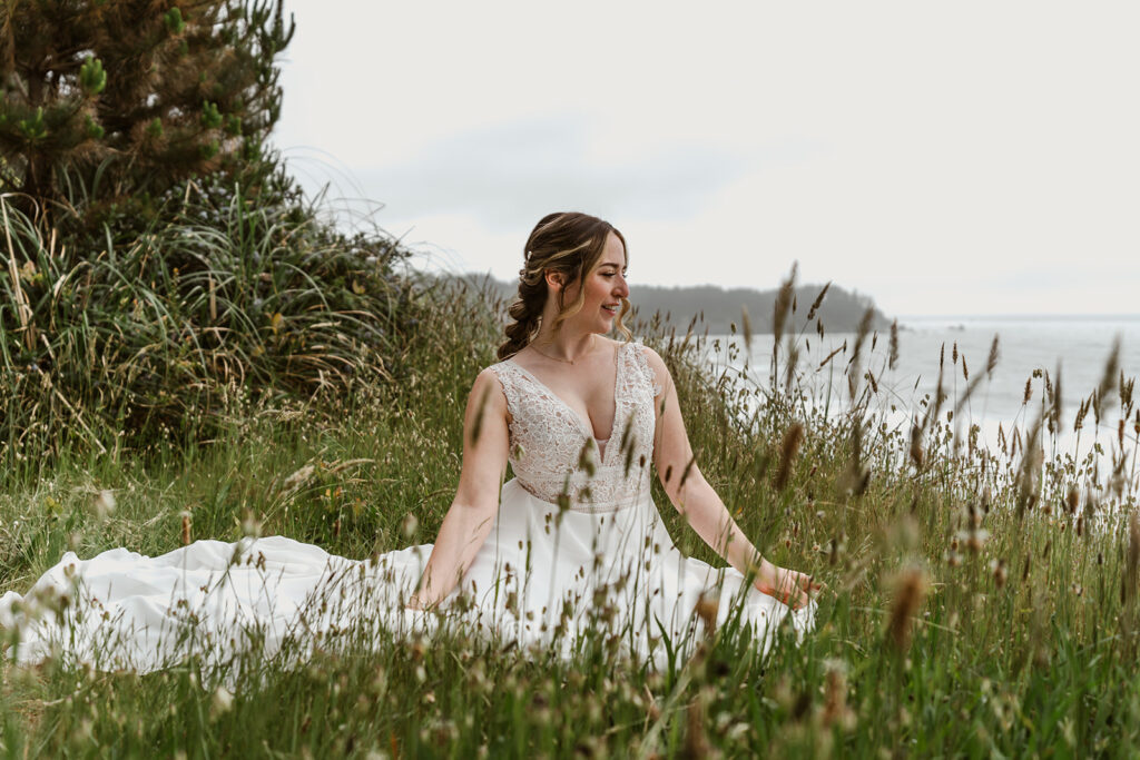 on the day of her forest wedding, a bride sits in the tall grass, gazing out at the sea. The train of the dress spills out behind her