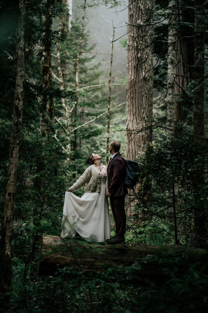 a couple in their wedding attire stand on a fallen tree as they explore during their forest wedding. They gaze up towards a pocket of light as they embrace.