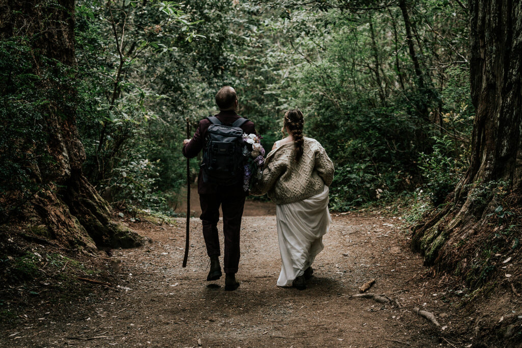 A joyful, horizontal shot from behind our bride and groom. They walk with excited skips in their steps. Our groom carries a walking staff and our bride has her dress gathered in her hands. 