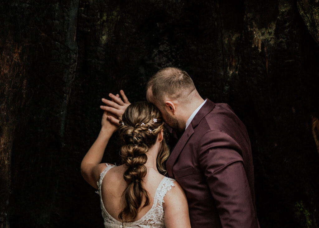 a couple in wedding attire press their heads together as they layer their hands on the bark of a Redwood tree. The whisper a quiet prayer as they begin their forest wedding. 