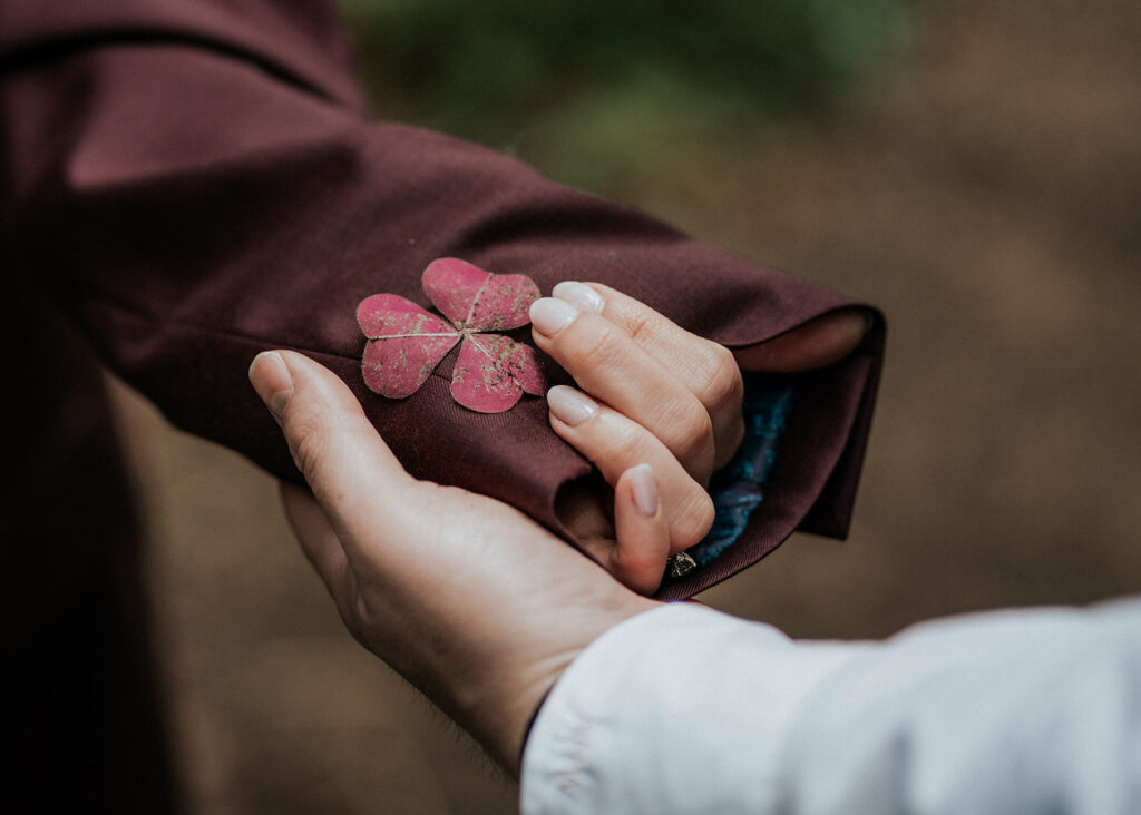 a wide, detailed shot of our couples hands. In our brides hands, we see that she is wearing her grooms jacket while holding a red clover. It matches the red of the clover almost perfectly. 