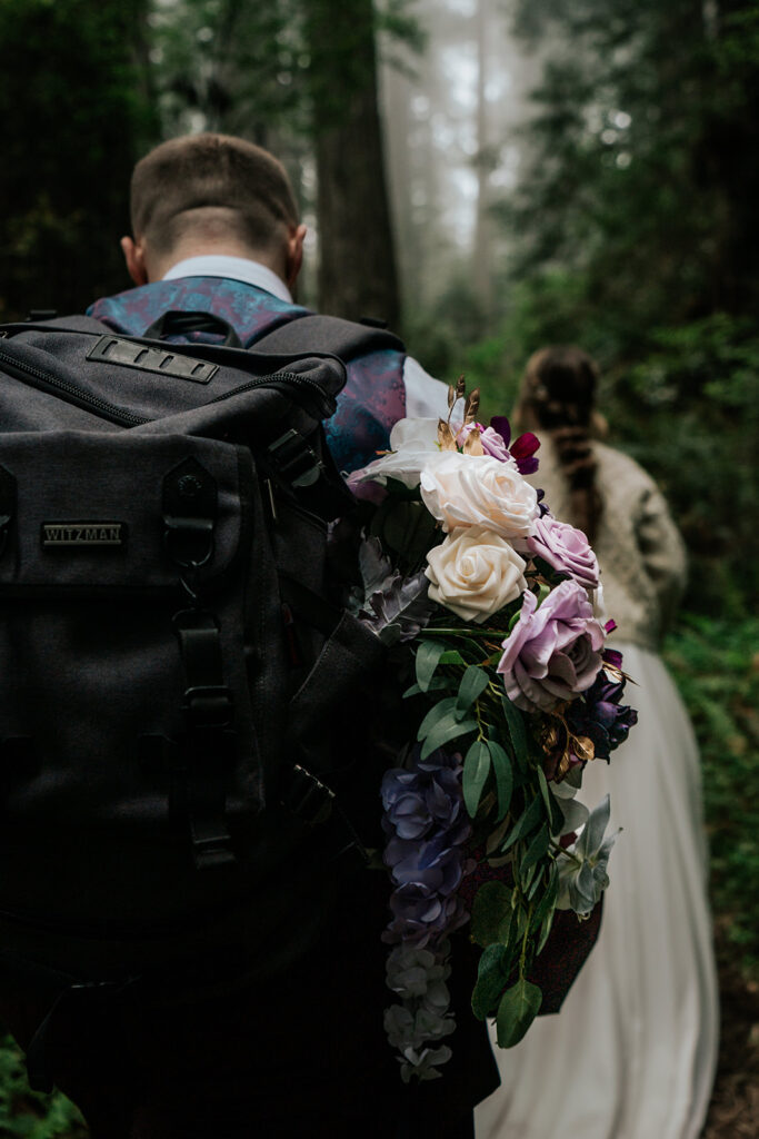 A shot from behind our couple as they walk to their forest wedding location. We see our bride walking in the front, our groom carries her bouquet in his backpack