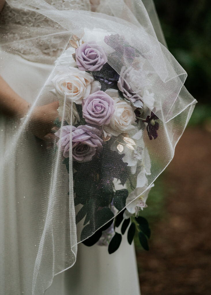 A sparkled veil cascades over the light purple and white bouquet held by our bride