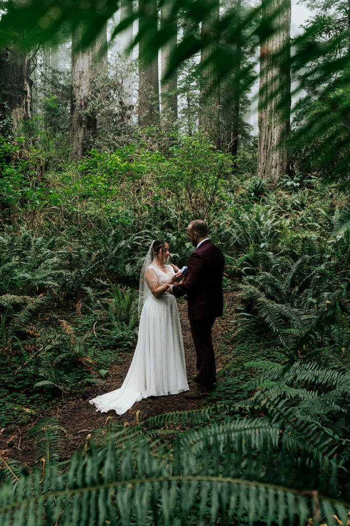 Surrounded and framed by deep, green ferns, we see our bride read her vows to her groom during their forest wedding.