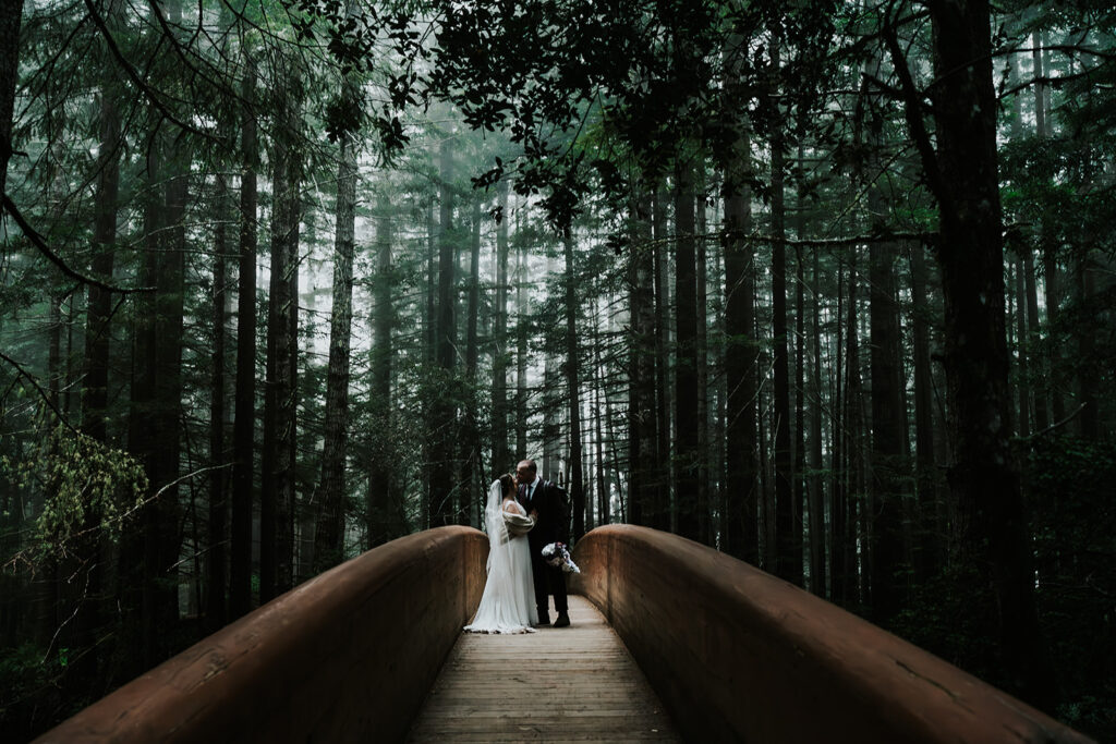 a bride and groom kiss on a bridge, surrounded by trees as they leave their forest wedding