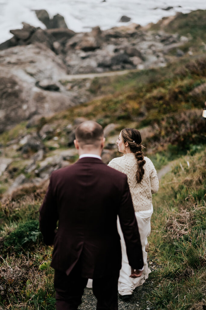 A bride and groom walk down a rocky trail to a rugged beach landscape 