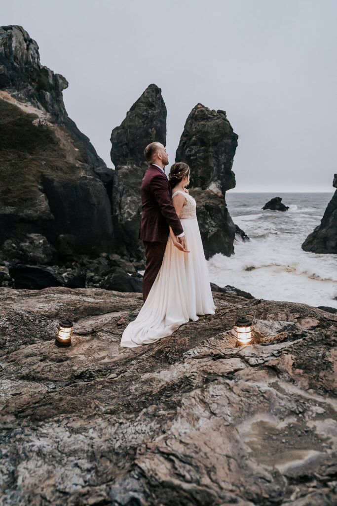 a bride and groom gaze out at the ocean surrounded by massive sea rocks 