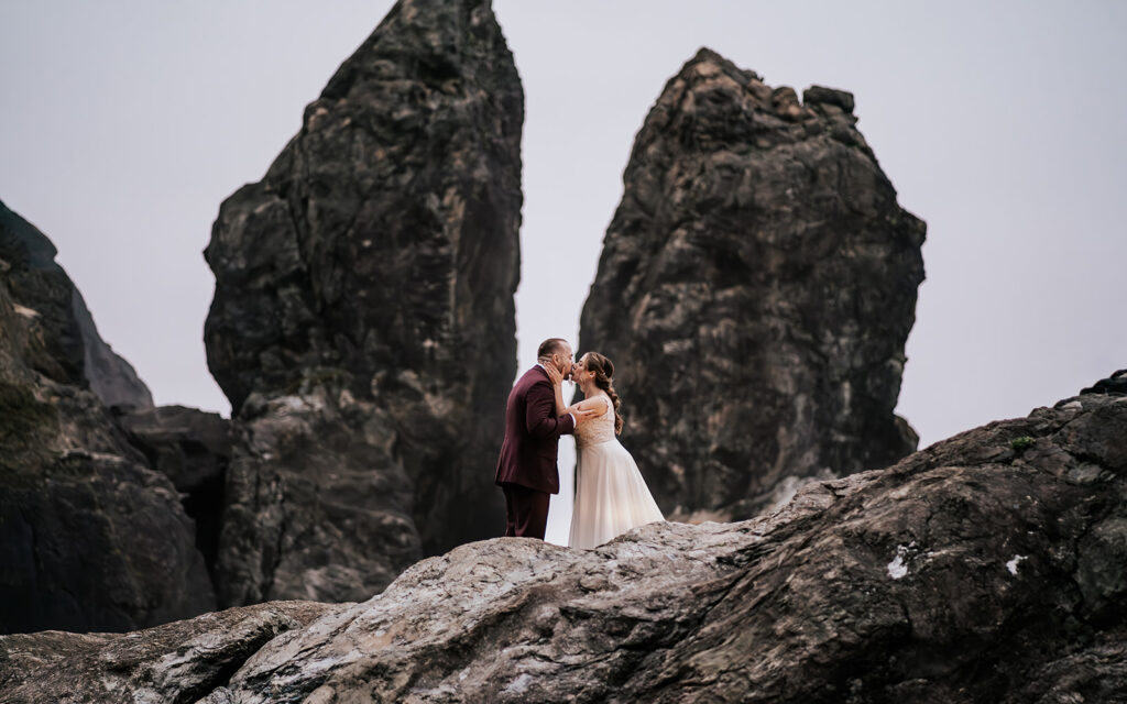 A bride and groom kiss in front of two huge vertical sea rocks