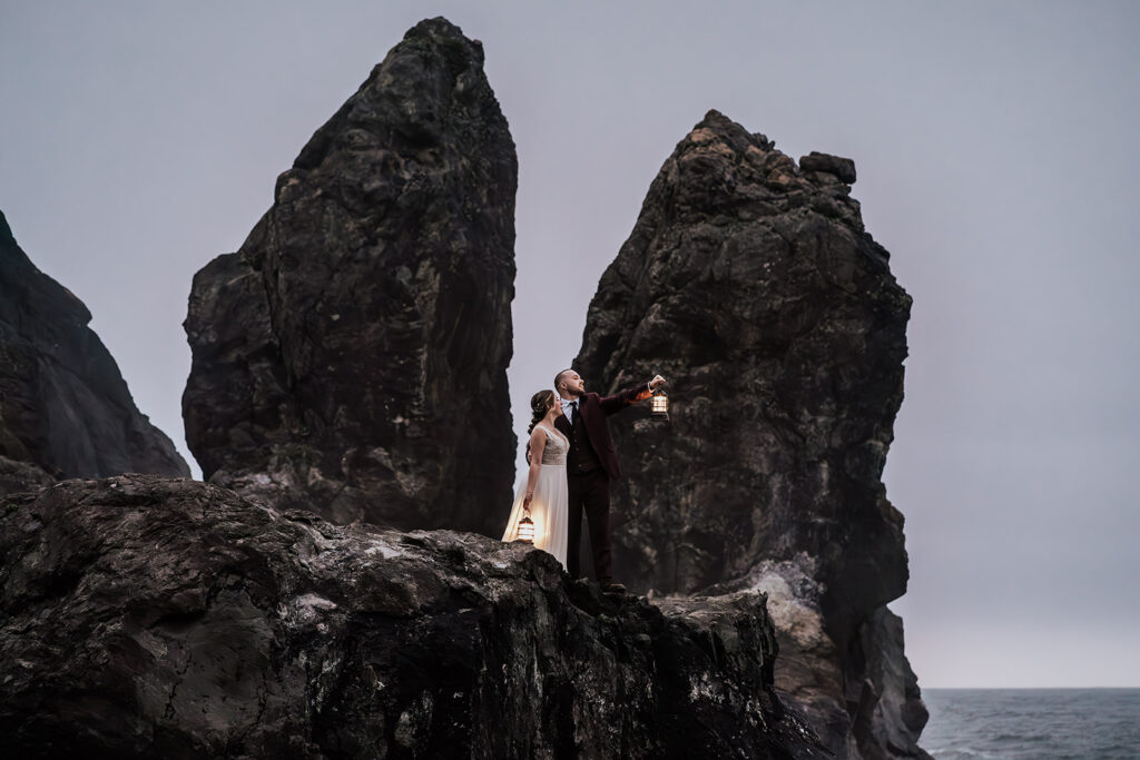 A couple in wedding attire stands on massive rocks on the beach. A calm ocean before them. It is dusk and the light is low, they hold lanterns high to give light.