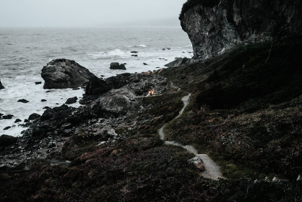 A wide landscape shot of a rocky shoreline. A gravel path creates a leading line to a couple in their wedding attire. It is Dusk and they hold lanterns to light their way through the landscape.