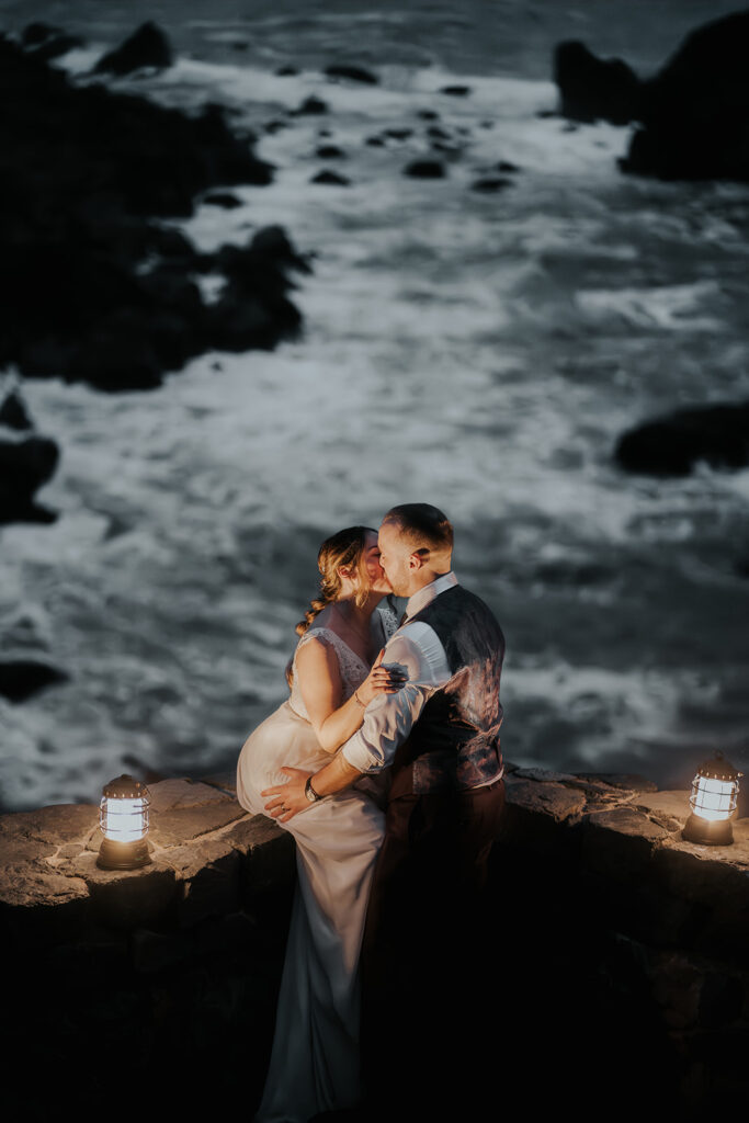 sitting on a stone wall, a bride and groom kiss by lantern light as the ocean water swirls beneath them, concluding their forest wedding day