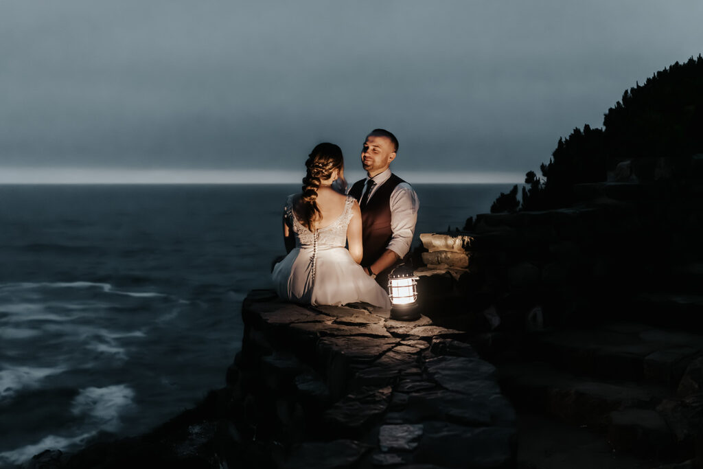 A bride sits on a stone wall gazing at her groom by lantern light at the end of their forest wedding day