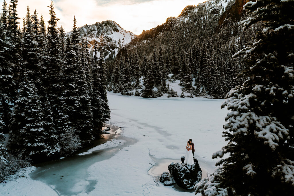 A bride and groom in wedding attire embrace on a frozen lake surrounded by mountains and pines during their backpacking wedding.  