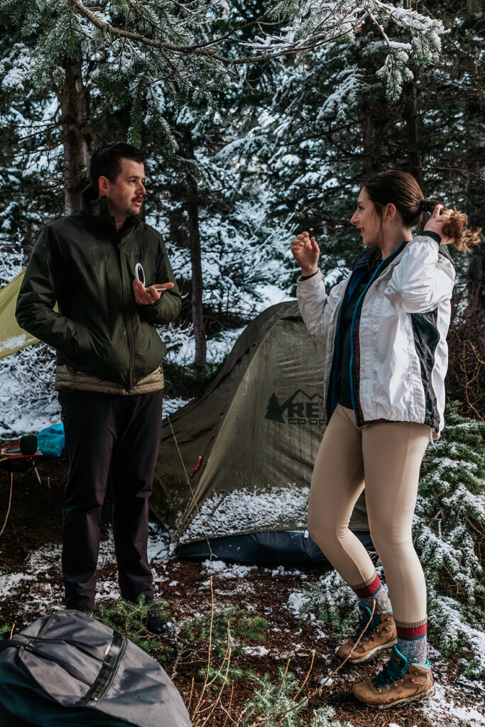 A bride and groom get ready for their backpacking wedding together. Both in warm layers, the groom holds a small mirror for his soon-to-be bride as she fixes her hair