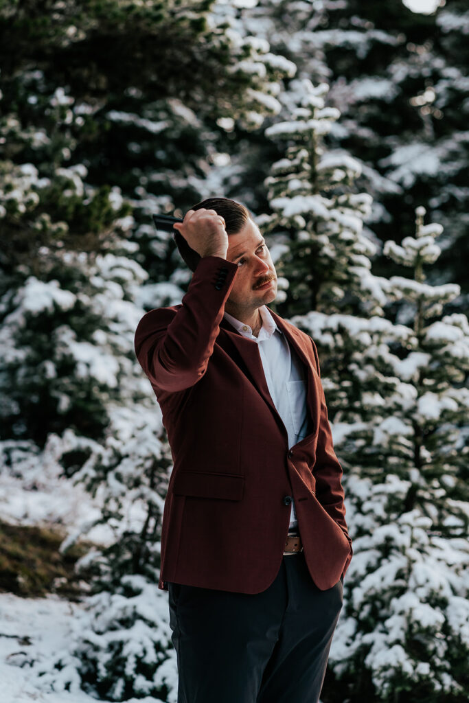 A groom, in his suit gets ready for their backpacking wedding by combing his hair while standing out in the snowy landscape, surrounded by pines