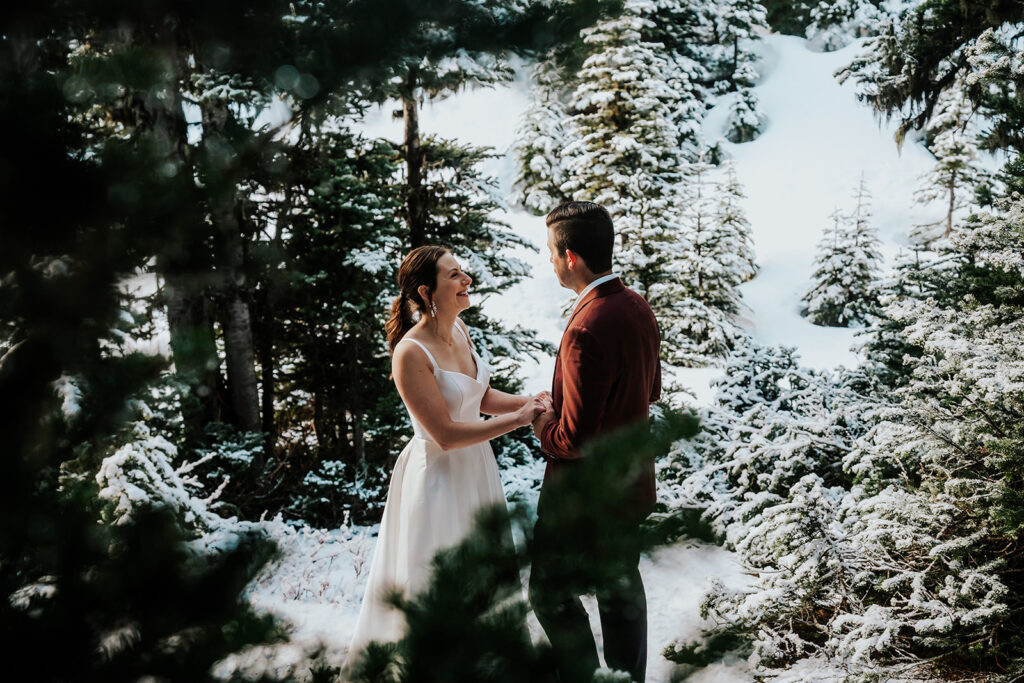 During their backpacking wedding a couple embraces between to pines. the landscape is blanketed with fresh snow. They smile lovingly at each other as they are framed by pine leaves