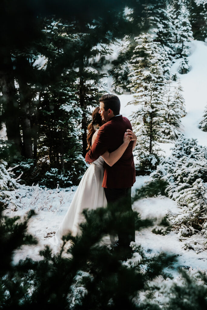 A bride and groom in wedding attire embrace framed by pine leaves during their snowy backpacking wedding.  