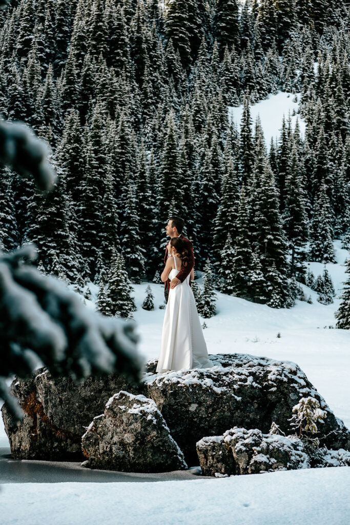 During their backpacking wedding a couple embraces, gazing at the landscape before them. the landscape is blanketed with fresh snow. Golden, morning light hits the faces of the mountains surrounding their ceremony.