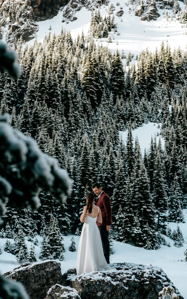 A bride and groom in wedding attire smile while standing on a rock surrounded by snowy mountains and pines during their backpacking wedding.  