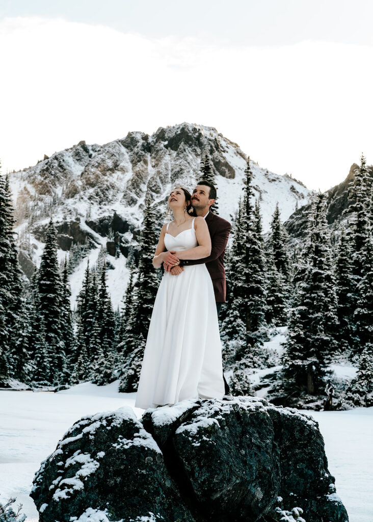 During their backpacking wedding a couple embraces, gazing at the landscape before them. the landscape is blanketed with fresh snow. Golden, morning light hits the faces of the mountains surrounding their ceremony.