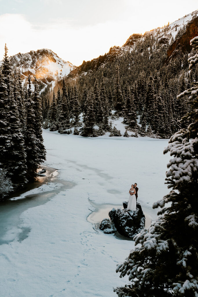 A bride and groom in wedding attire embrace on a frozen lake surrounded by mountains and pines during their backpacking wedding.  