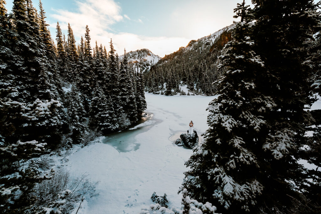 A bride and groom in wedding attire embrace on a frozen lake surrounded by mountains and pines during their backpacking wedding.  