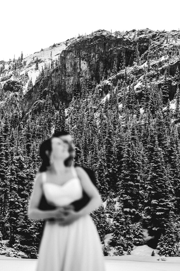 A blurry shot of a bride and groom in wedding attire embracing and looking to the sky. The landscape behind them is in focus and consists of mountains and pines 