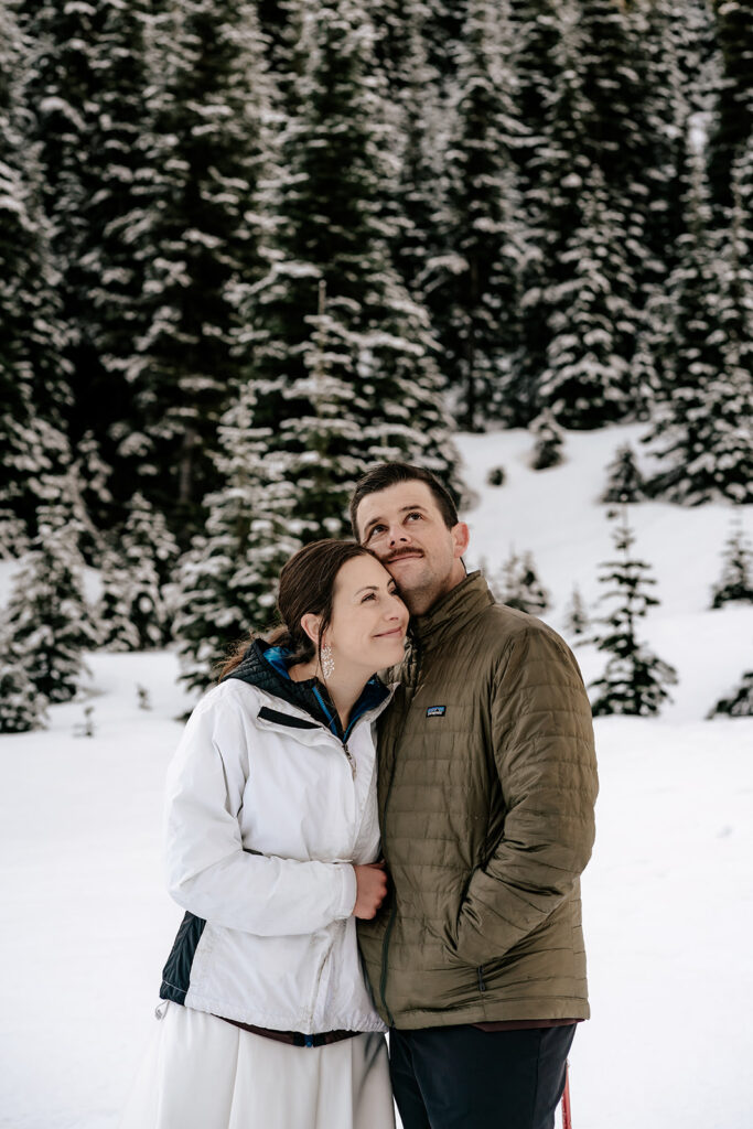 A bride and groom in wedding attire explore a snowy landscape with mountains and pines during their backpacking wedding. 