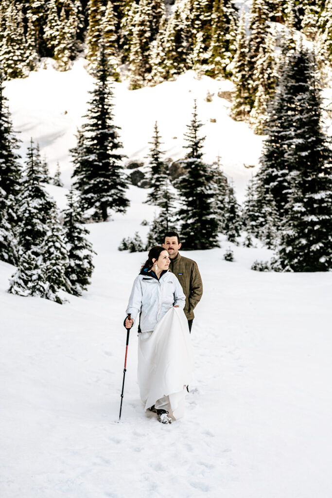 A bride and groom in wedding attire explore a snowy landscape with mountains and pines during their backpacking wedding. 