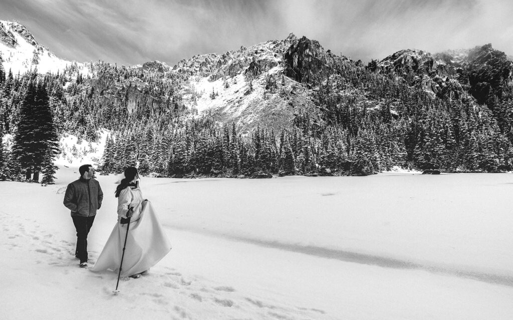 A black and white landscape image of a bride and groom in wedding attire explore a snowy landscape with mountains and pines during their backpacking wedding. 