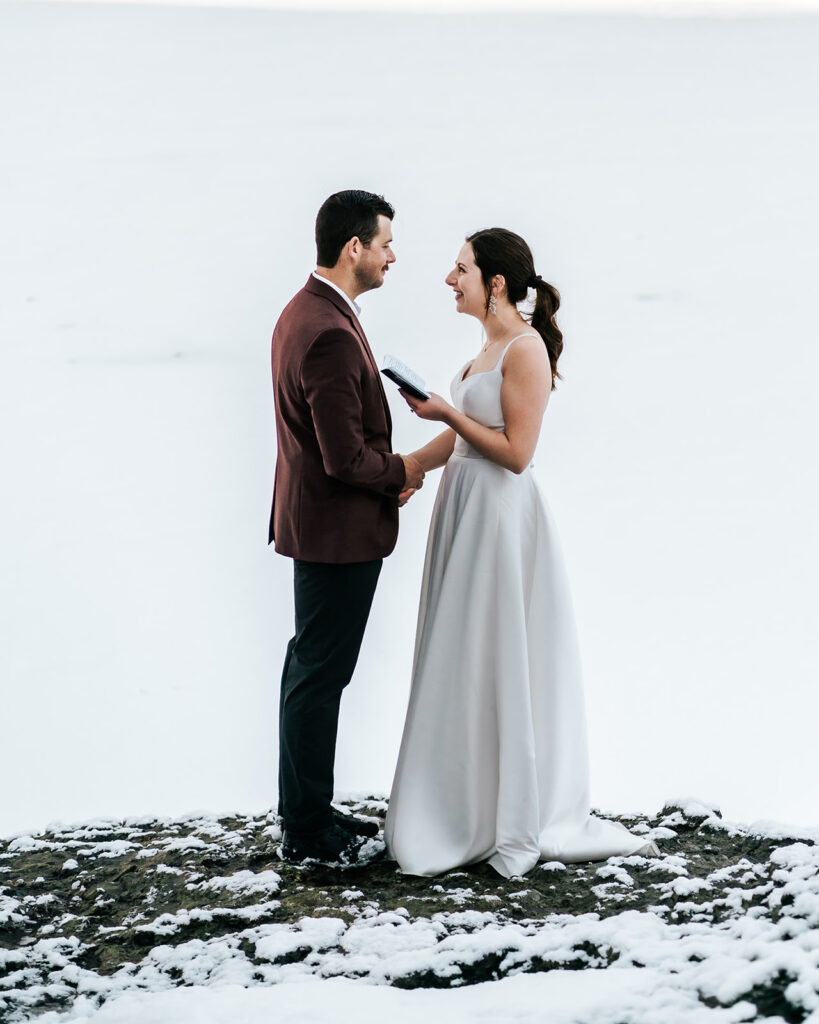 A bride and groom holding hands. In the brides free hand, she holds her vow book open as she reads her vows to her groom during their backpacking wedding