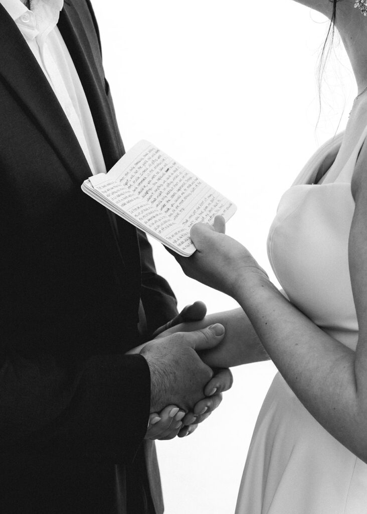 a close up, black and white image of a bride and groom holding hands. In the brides free hand, she holds her vow book open as she reads her vows to her groom during their backpacking wedding