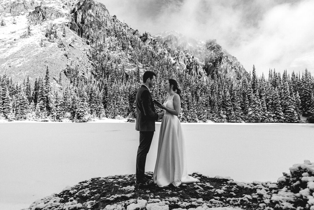 A black and white image of bride and groom holding hands. In the brides free hand, she holds her vow book open as she reads her vows to her groom during their backpacking wedding. they stand on a rock surrounded by mountains 