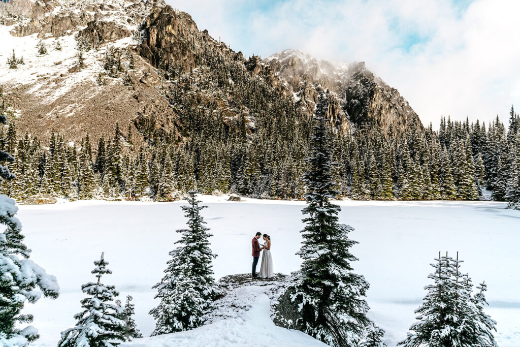 During their backpacking wedding a couple embraces between to pines. the landscape is blanketed with fresh snow. Golden, morning light hits the faces of the mountains surrounding their ceremony.