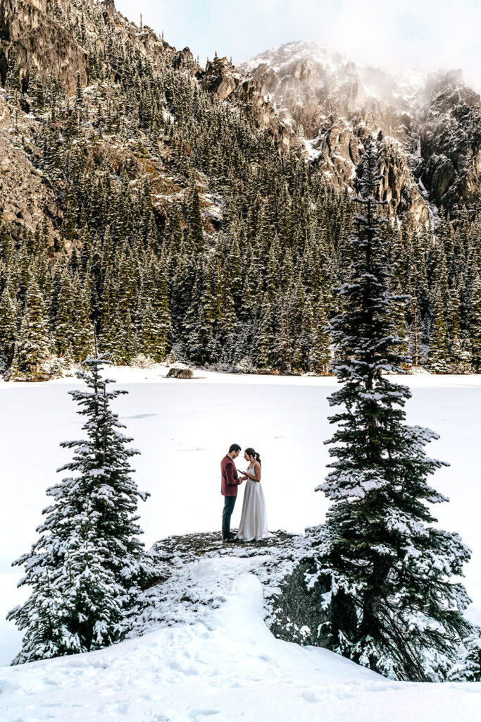 During their backpacking wedding a couple embraces between to pines. the landscape is blanketed with fresh snow. Golden, morning light hits the faces of the mountains surrounding their ceremony.