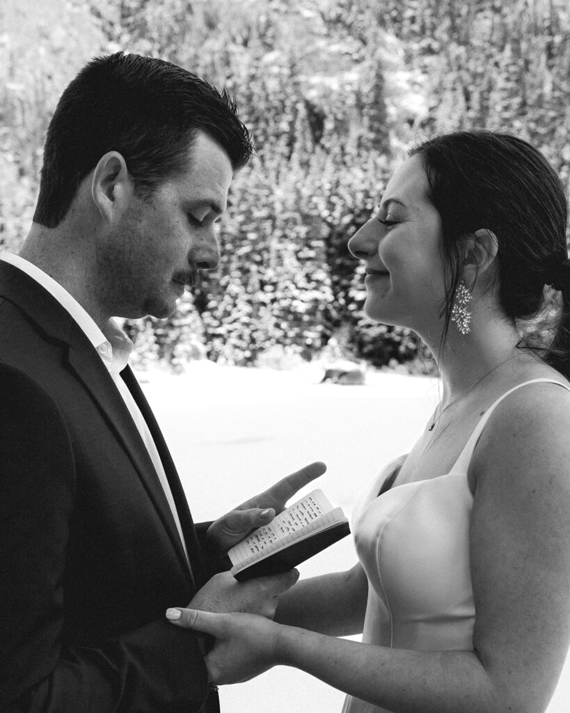 a close up, black and white image of a bride and groom holding hands. In the grooms free hand, he holds his vow book open as he reads his vows to his bride during their backpacking wedding