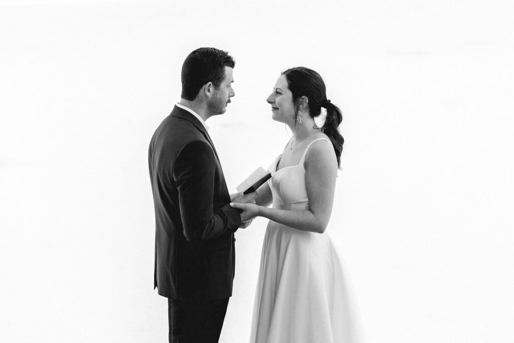 a wide, black and white image of a bride and groom holding hands. In the grooms free hand, he holds his vow book open as he reads his vows to his bride during their backpacking wedding