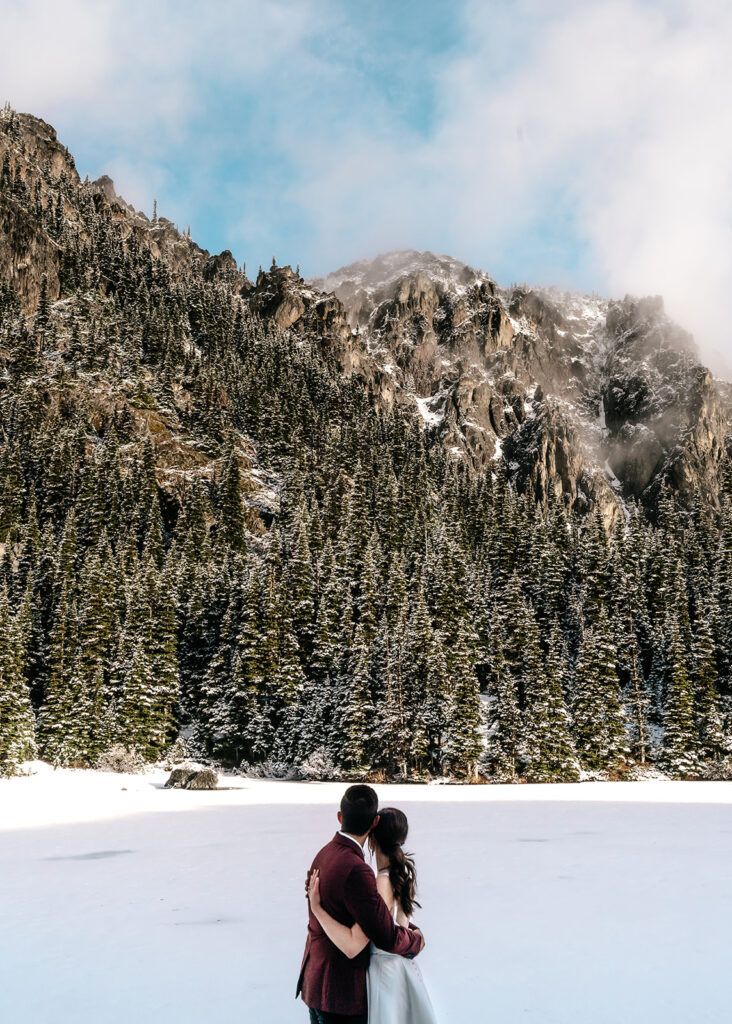 During their backpacking wedding a couple embraces, gazing at the landscape before them. the landscape is blanketed with fresh snow. Golden, morning light hits the faces of the mountains surrounding their ceremony.