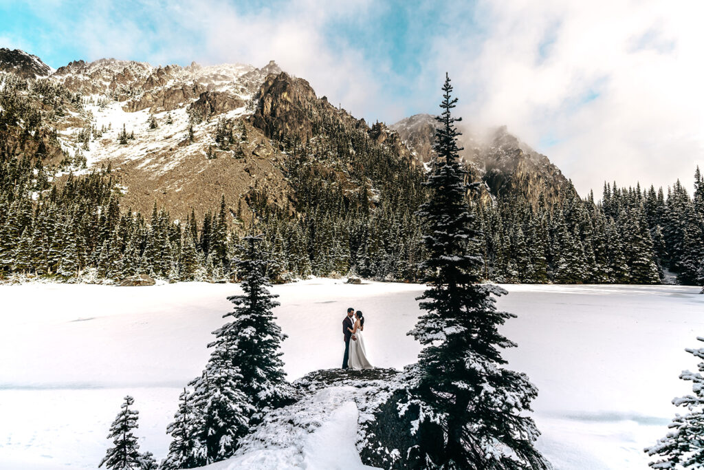 During their backpacking wedding a couple embraces between to pines. the landscape is blanketed with fresh snow. Golden, morning light hits the faces of the mountains surrounding their ceremony.