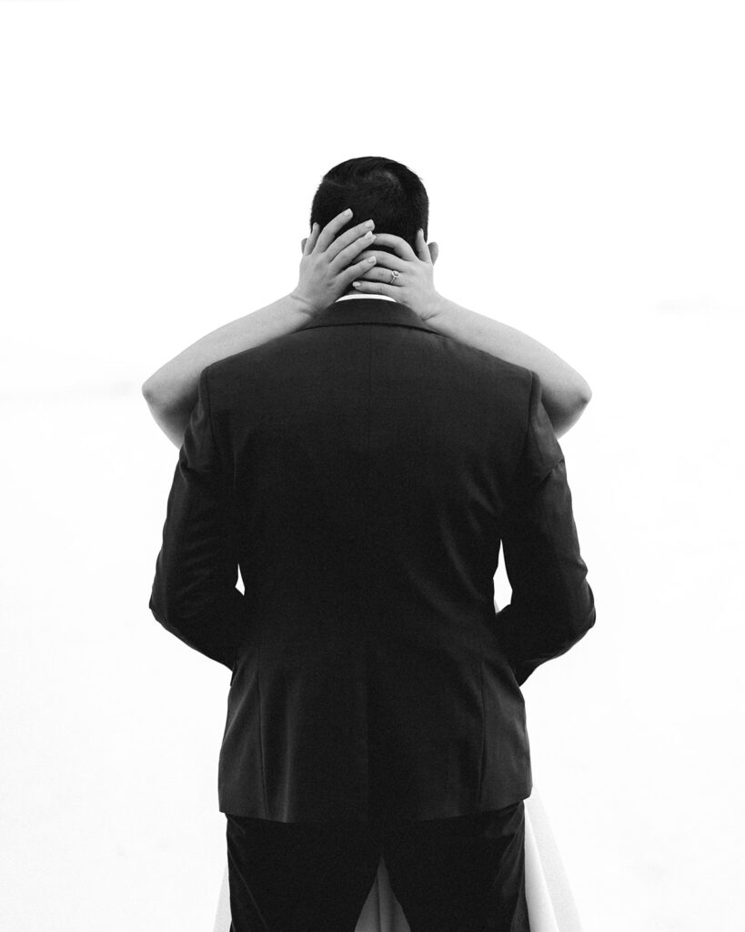 a black and white image of the bride and groom embracing during their backpacking wedding. Shot with the grooms back to the camera we see only the hands of his bride wrapped around his shoulders