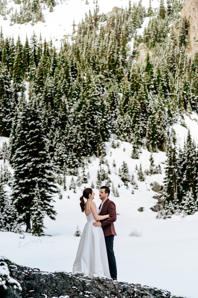 During their backpacking wedding a couple embraces, gazing at the landscape before them. the landscape is blanketed with fresh snow. Golden, morning light hits the faces of the mountains surrounding their ceremony.