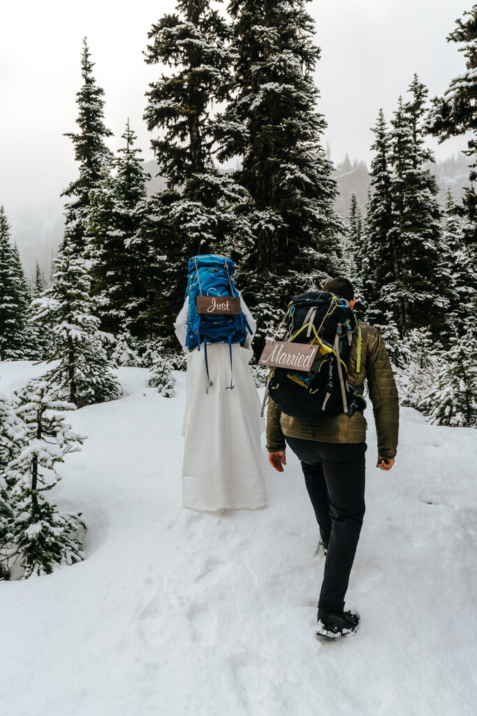 A bride and groom in wedding attire and backpacking gear explore a snowy landscape with mountains and pines during their backpacking wedding. wooden signs reading "just married" hang from their packs. 