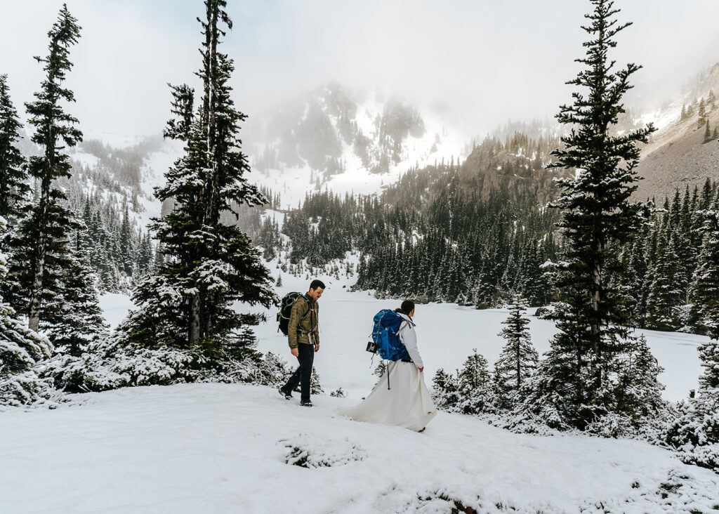 A bride and groom in wedding attire and backpacking gear explore a snowy landscape with mountains and pines during their backpacking wedding. 