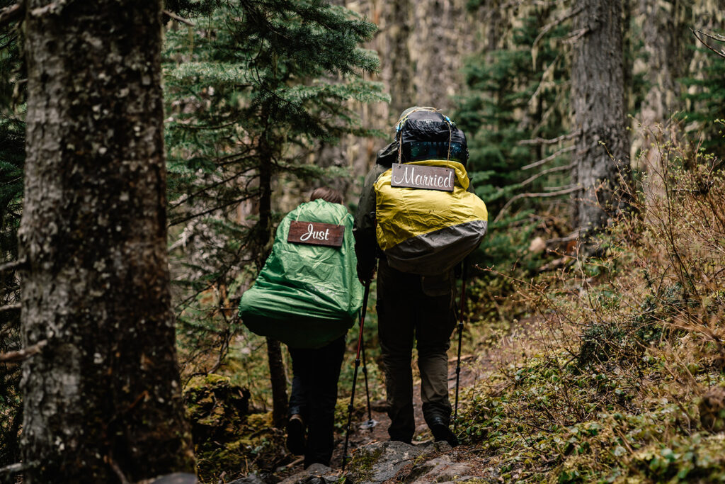 a couple in backpacking attire and backpacking bags walk side by side as they explore during their backpacking wedding. Wooden signs reading "just married" hang from their packs