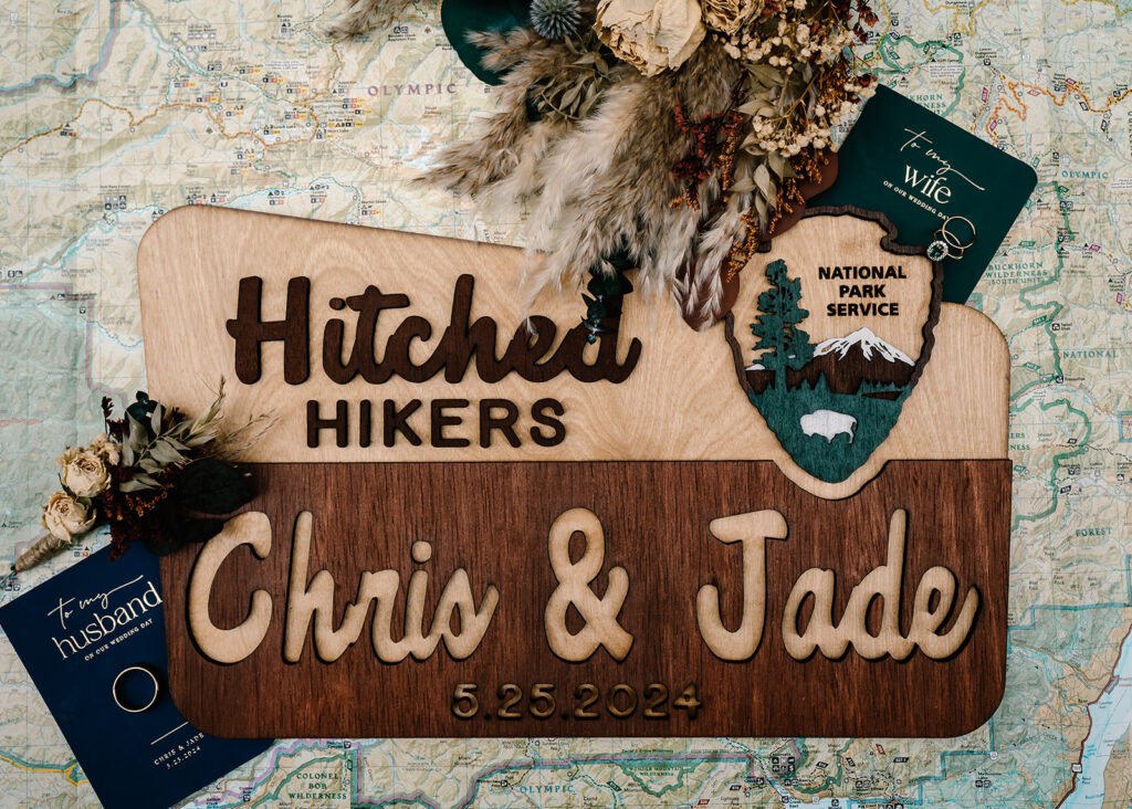 details from this backpacking wedding arranged on a map of the park and include a wooden sign in national park style that says "hitched hikers" personalized with their names and date. Included in this detail shot are their blue and green vow books and dried florals