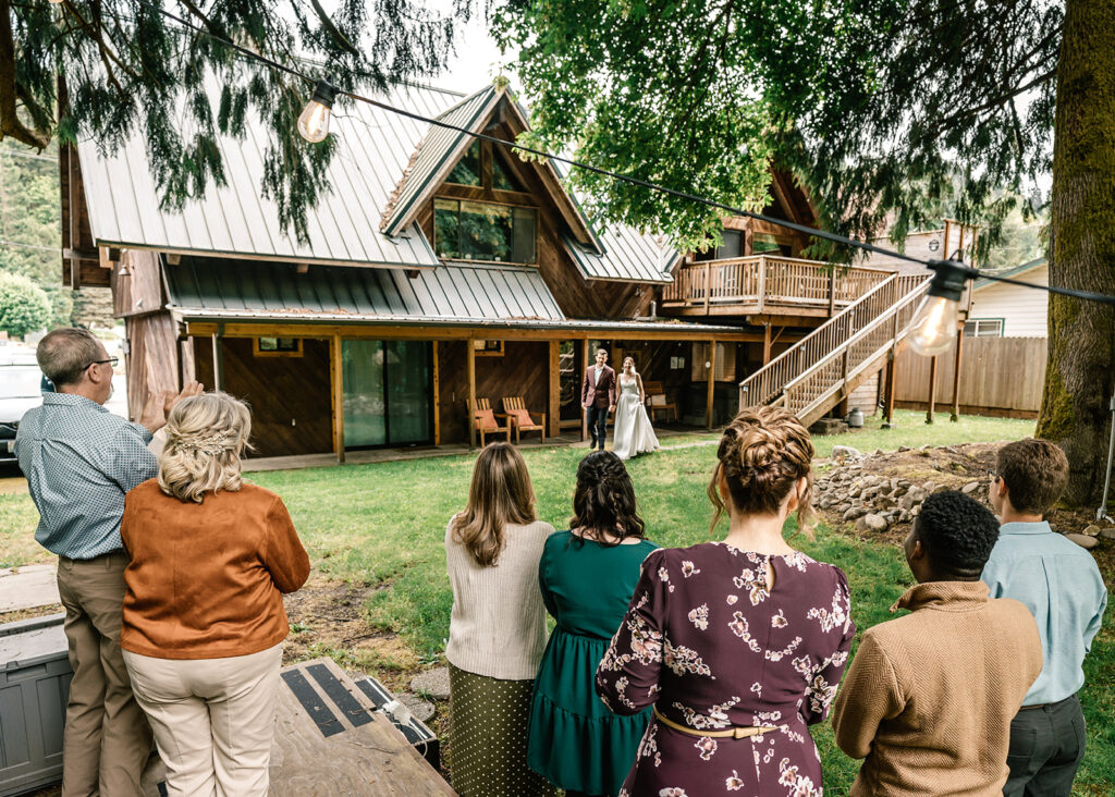 taken from the perspective of their guests. during their backpacking wedding, a couple walks outside their cabin in their wedding attire. They smile widely as they greet their guests for the first time in their wedding attire