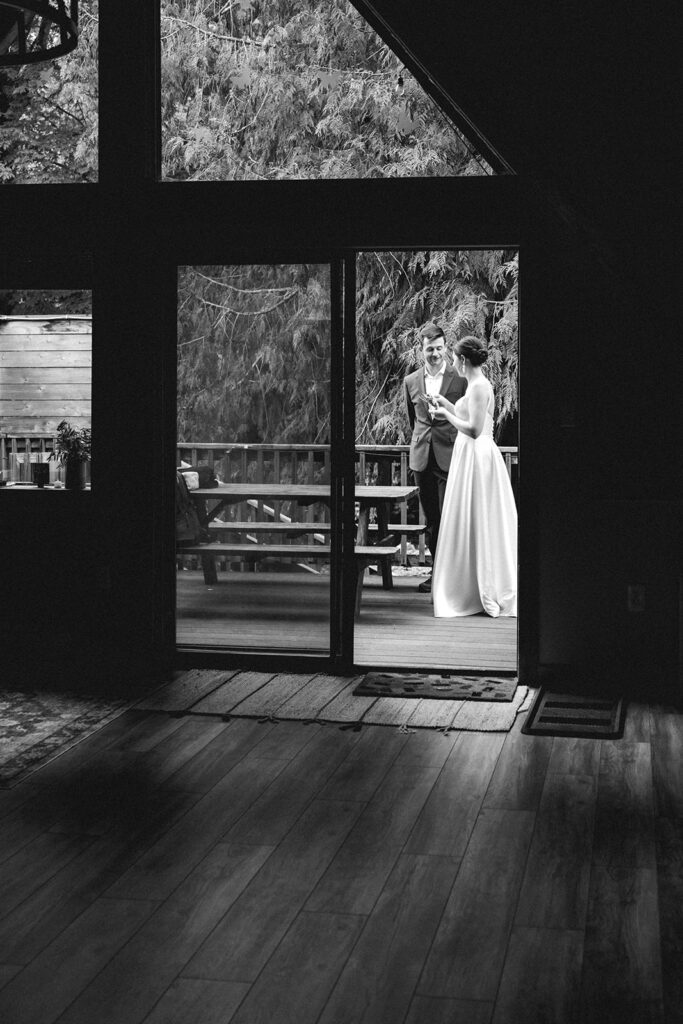 During their backpacking wedding, a bride and groom in their wedding attire, stand outside of their lodging. they are framed by the architecture of the A-frame style doorway.