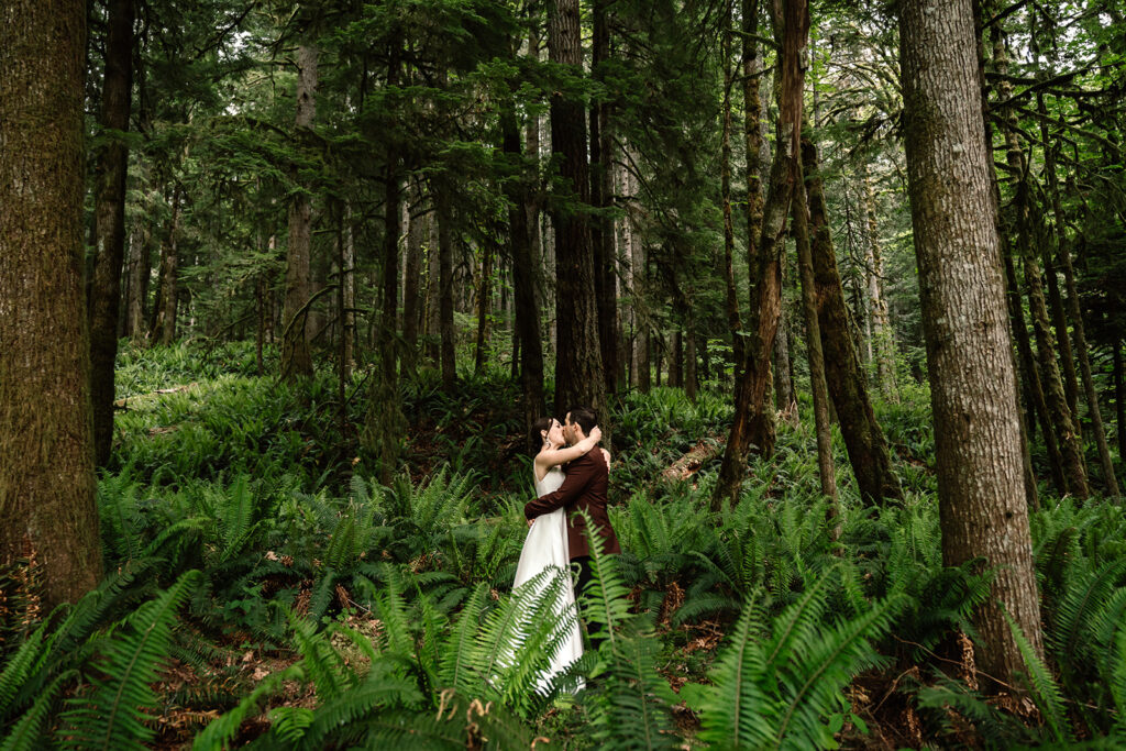 during their backpacking wedding, a bride and groom kiss and embrace in a lush patch of forest, they are framed by ferns and trees. 