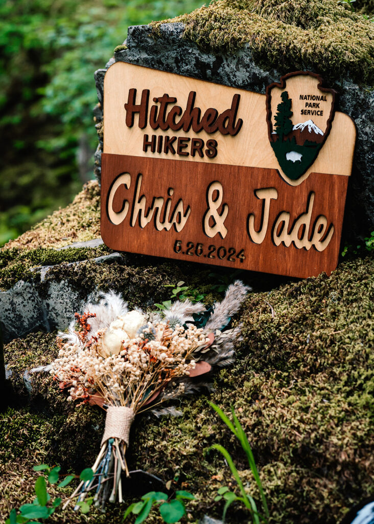 details from this backpacking wedding including a national park style sign reading "hitched hikers", customized with their names and wedding date. The sign and the brides bouquet all rest on a mossy rock