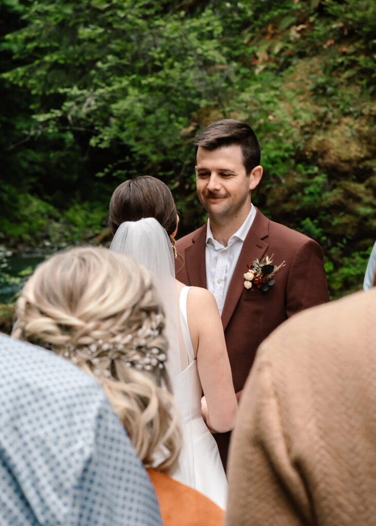 shot over the shoulder of their guests, a groom smiles as he gazes lovingly at his bride during their backpacking wedding